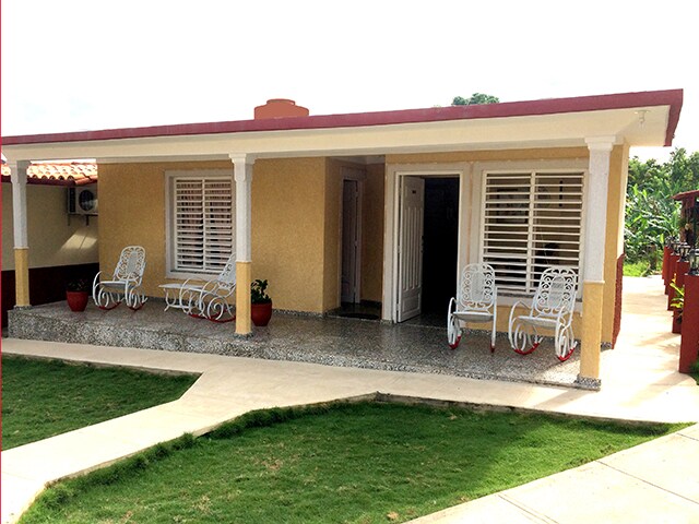 A charming single-story house with a covered porch features white wicker chairs arranged on a stone pathway. Large windows showcase natural light, and well-maintained greenery surrounds the entrance, creating a welcoming and bright exterior. The simple design exudes tranquility.