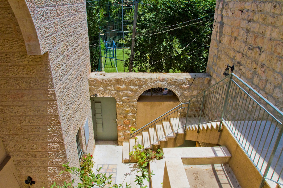 A stone staircase is visible, leading down to a lower level, flanked by natural greenery. The area features textured stone walls, and an arched opening provides access to a door at the bottom. Soft light illuminates the space, enhancing its tranquil ambiance.