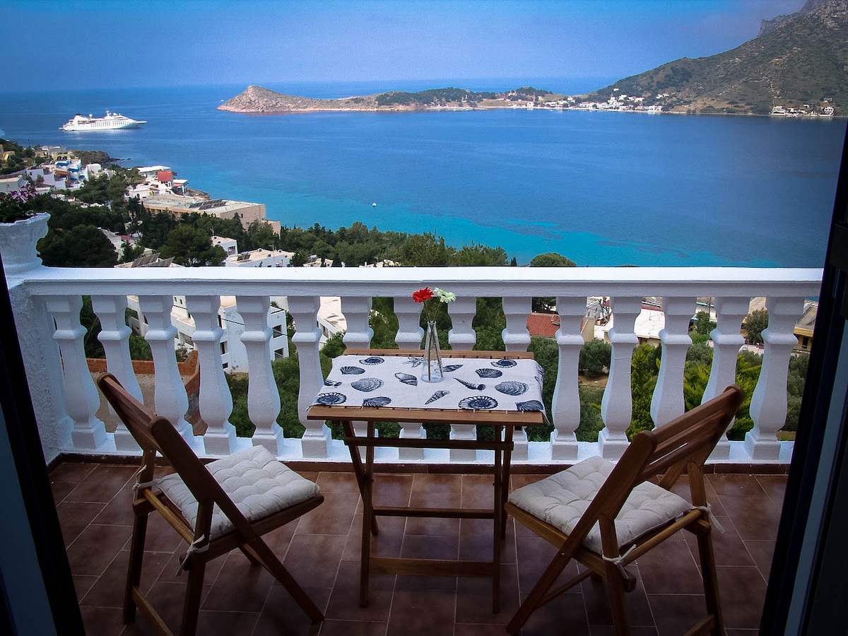 A spacious outdoor seating area is depicted, featuring a small table adorned with a vase and flowers. Two wooden chairs and a scenic view of the coastline and the island of Telendos can be seen in the background, creating an inviting space to relax.