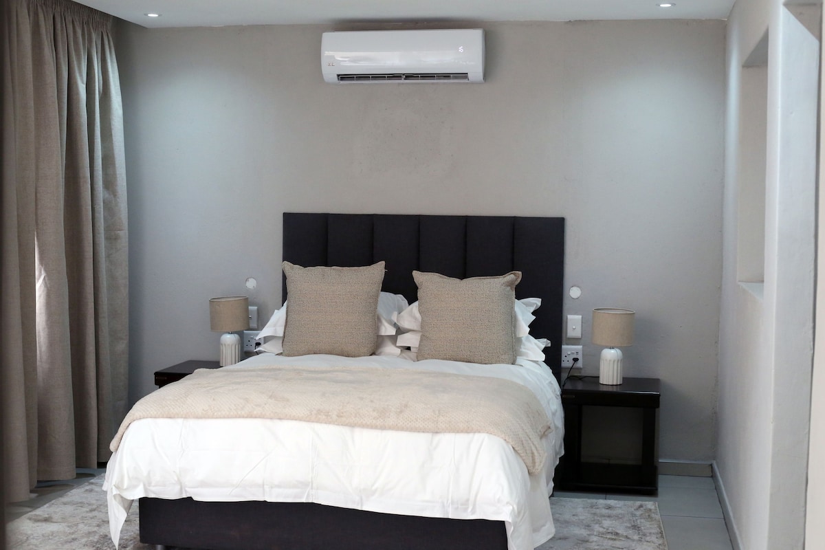A tranquil bedroom is presented with a dark upholstered headboard and two large cushions. Nightstands with lamps are positioned on either side of the spacious bed, and natural light fills the room through flowing curtains. An air conditioning unit is mounted above the bed.