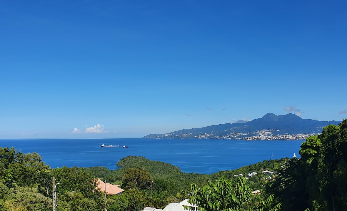 A panoramic view of the bay showcases the tranquil blue waters of Fort de France, framed by lush greenery. Sailboats can be seen gently bobbing in the distance against a clear blue sky, providing a serene backdrop to the coastal landscape.