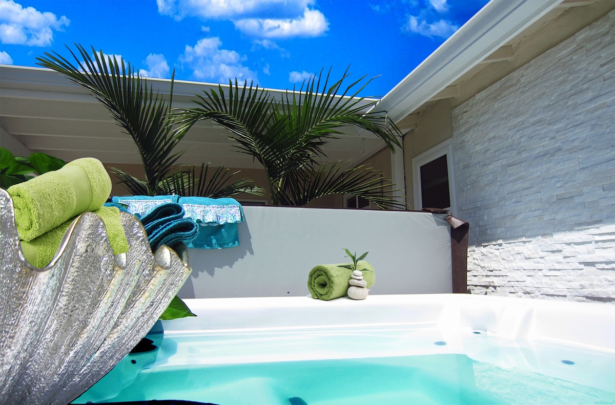 A sparkling hot tub is surrounded by lush greenery, with soft towels neatly placed nearby. The clear blue sky can be seen above, enhancing the serene atmosphere. Smooth stones and a small plant add a natural touch to the inviting space.