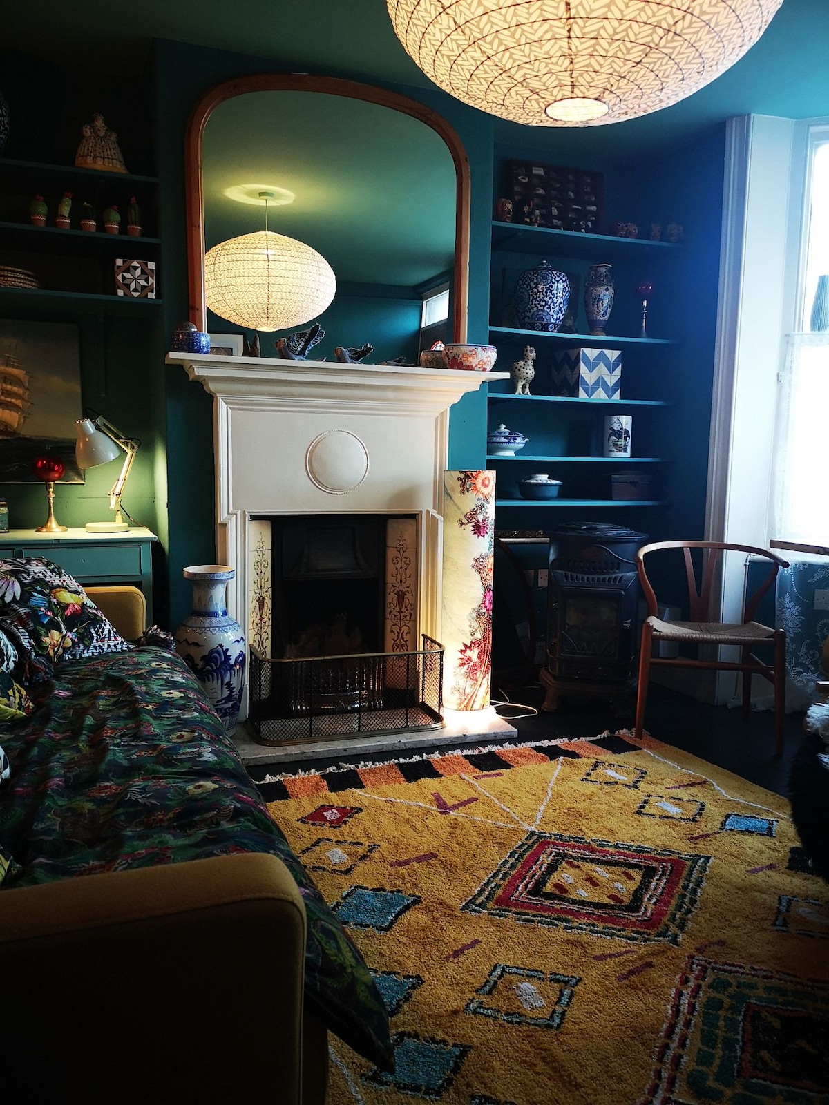 A colorful living area features a vibrant rug with geometric patterns covering the floor. A decorative fireplace serves as a focal point, surrounded by shelves displaying various unique items. Soft lighting is provided by a statement pendant lamp hanging above.