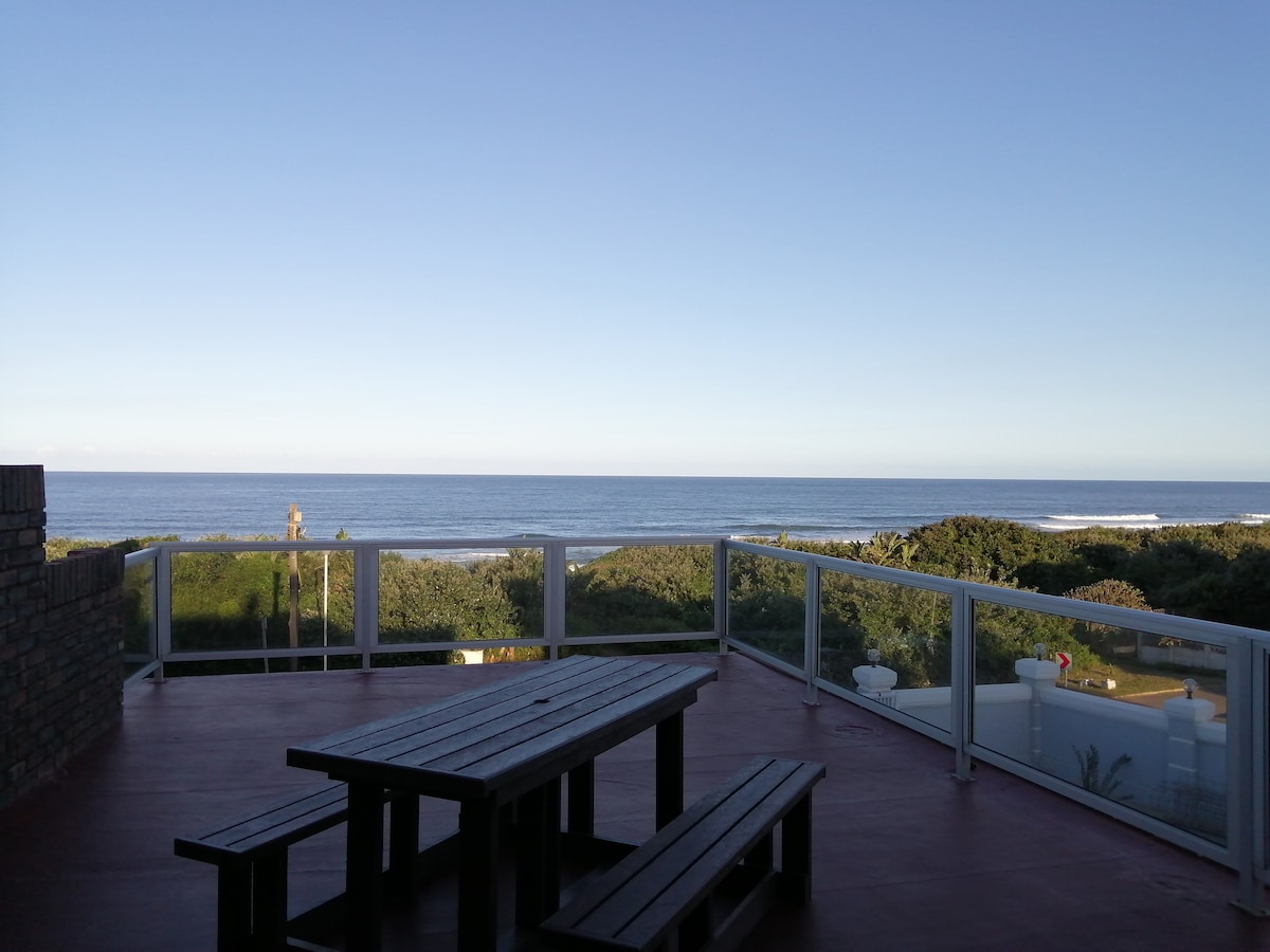 An outdoor deck area features a wooden picnic table, offering an unobstructed view of the ocean and surrounding greenery. Gentle waves can be seen along the horizon under a clear blue sky, creating a serene coastal environment.