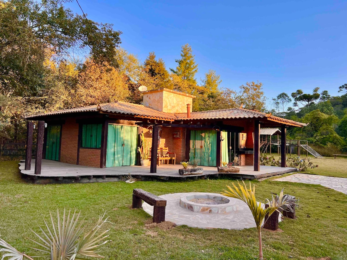 A one-story house with a rustic design features large green shutters and a spacious porch. Surrounding areas include a well-maintained lawn and a fire pit, creating a comfortable and inviting outdoor space. Nearby trees and a playground add to the serene setting.