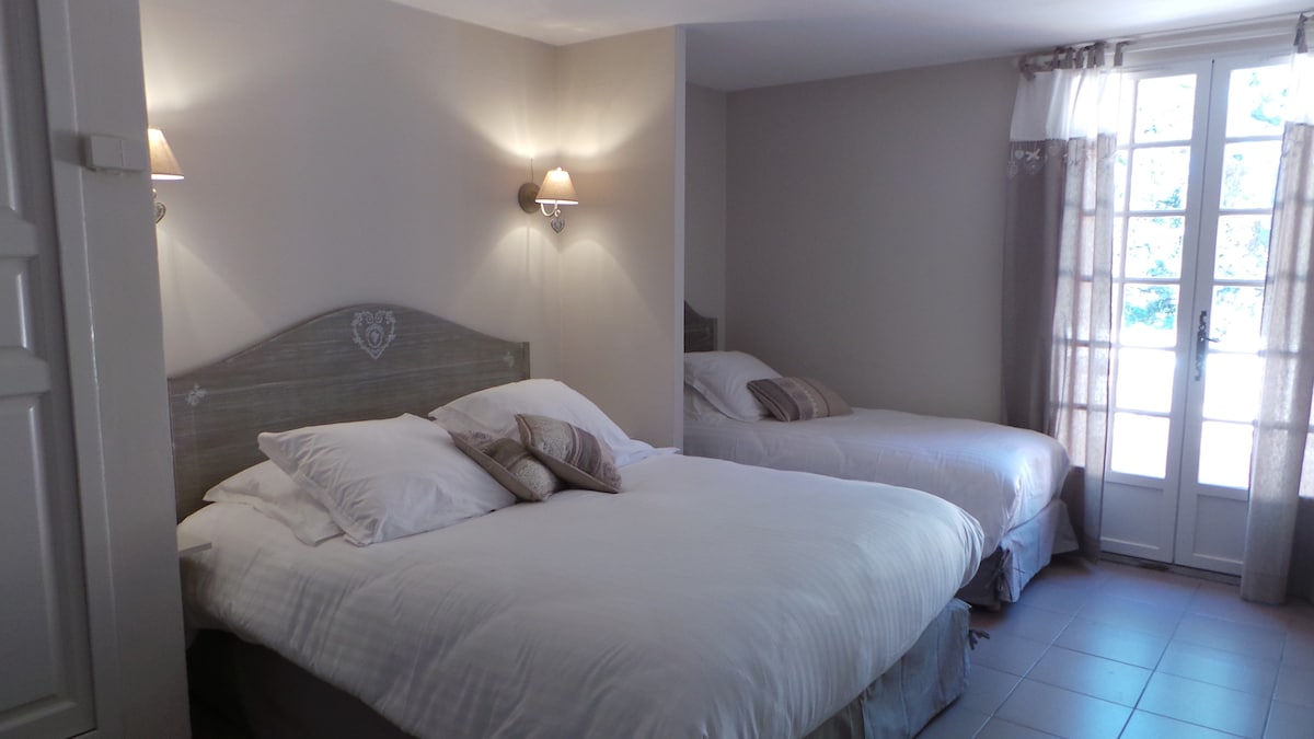 A comfortable guest room is presented with a spacious double bed and a single bed, both adorned with soft linens and decorative pillows. Natural light enters through a set of double doors, which open to an exterior area, enhancing the room's inviting ambiance.