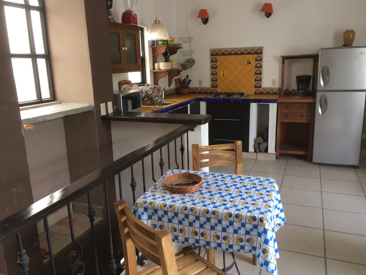 A brightly lit kitchen area features a blue and white checked tablecloth covering a wooden table, complemented by two wooden chairs. The kitchen counter showcases vibrant yellow tiles, and modern appliances, including a stainless steel refrigerator, are present in the space.