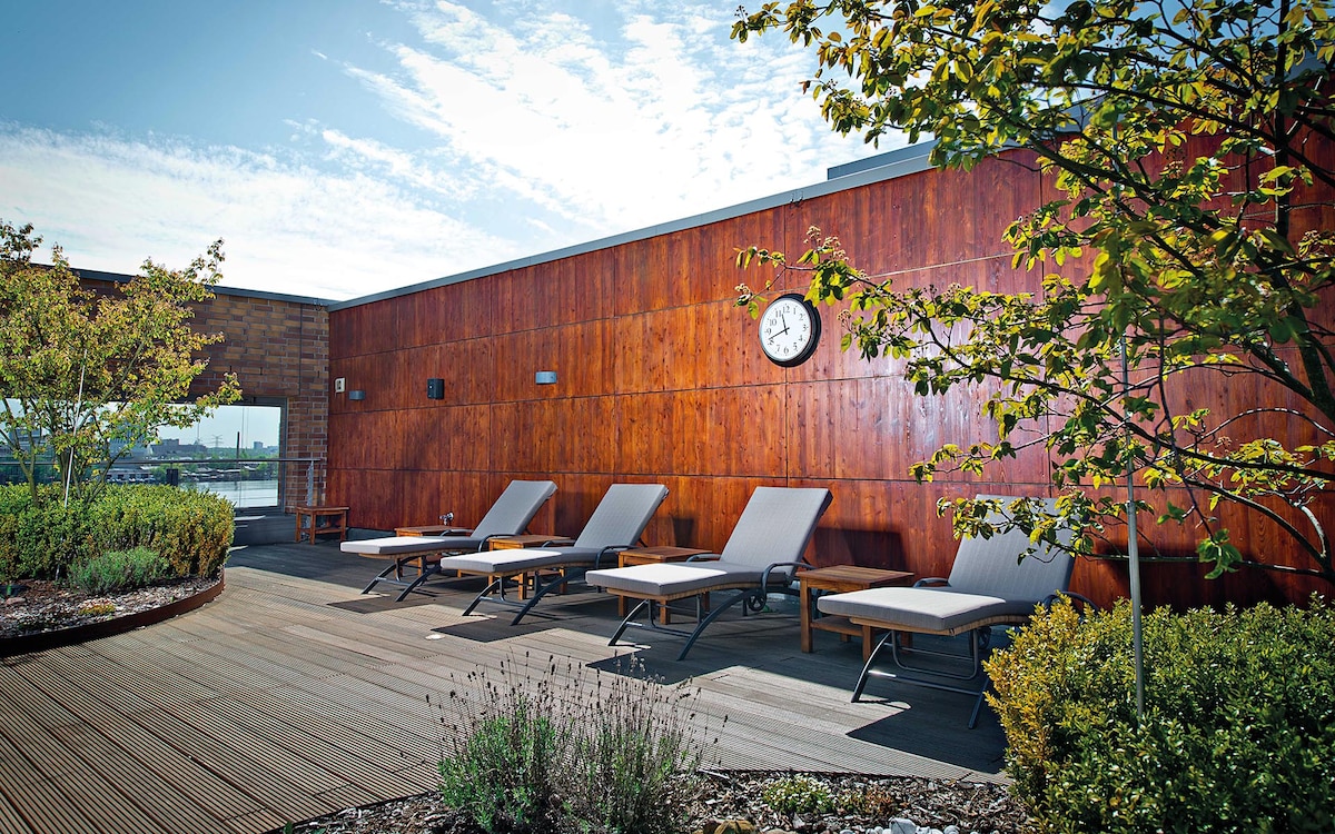 An outdoor space features several lounge chairs arranged on a wooden deck, surrounded by greenery and flowering plants. A wooden wall provides a warm backdrop, while a clock is mounted for easy visibility. The sky is clear and illuminated by natural light.