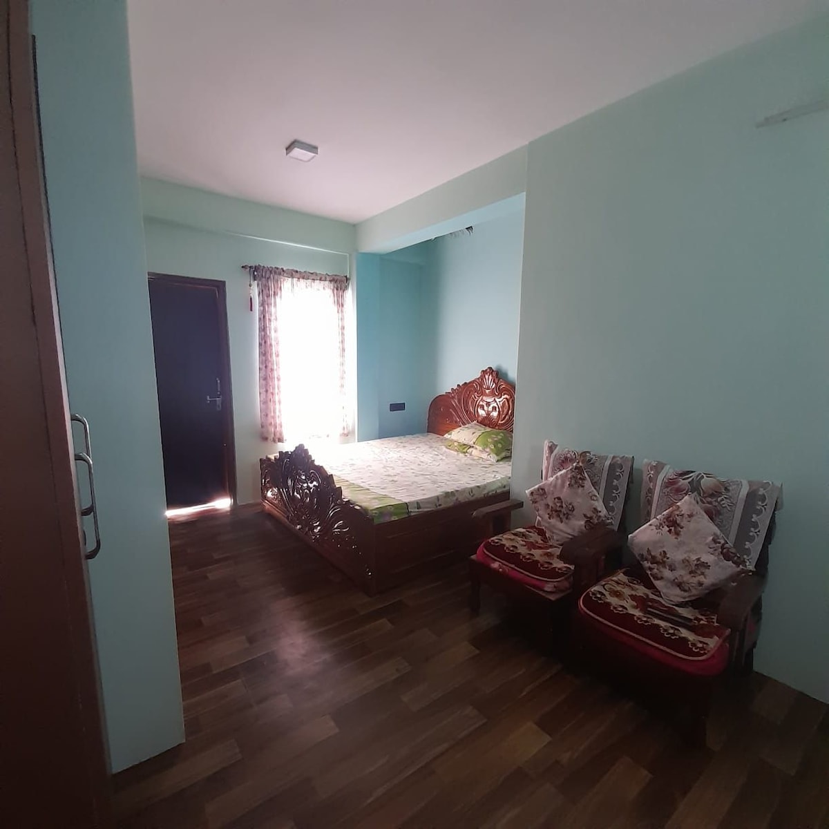 A well-lit sleeping area features a spacious bed with a decorative headboard against a light blue wall. Two wooden chairs, adorned with floral cushions, are situated next to a window dressed with sheer curtains, enhancing the room's brightness.