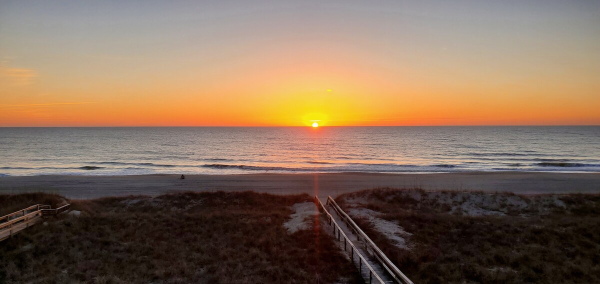 A stunning view of the ocean at sunrise is captured, showcasing vibrant hues of orange and yellow blending into the blue sea. The beach is visible in the forefront, framed by grasses and a wooden walkway leading toward the water.