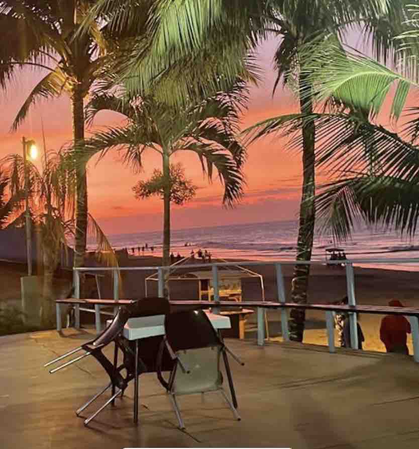 A deck features two chairs facing a vibrant sunset over the beach, with silhouettes of people walking along the shoreline. Lush palm trees frame the scene, while soft lighting illuminates the area, enhancing the connection to the ocean.