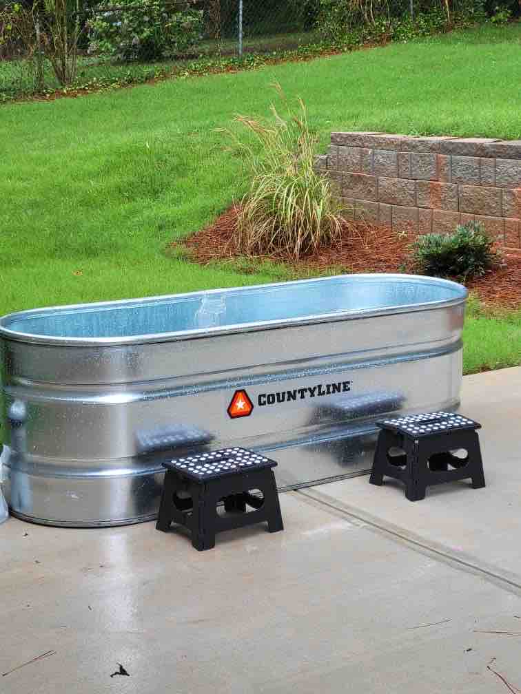 A metal livestock water trough is positioned on a concrete surface, accompanied by two small black stools. Lush green grass surrounds the area, contributing to a natural setting. The trough features the brand name 'COUNTYLINE' prominently displayed on one side.