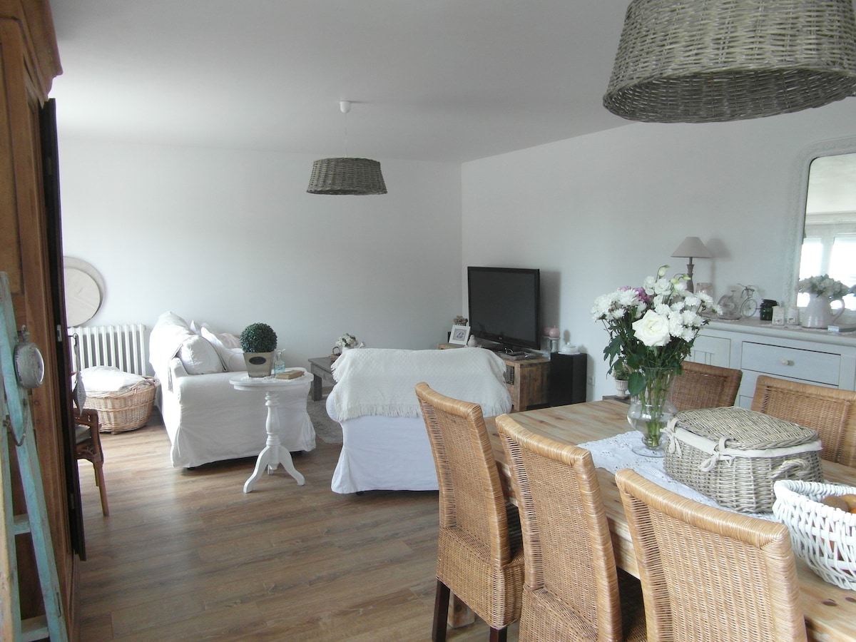 A bright living area features a wooden dining table surrounded by woven chairs. A cozy seating arrangement is positioned nearby, with a large television on display. Decorative elements include a basket and a floral arrangement, all set against light-colored walls and warm wood flooring.