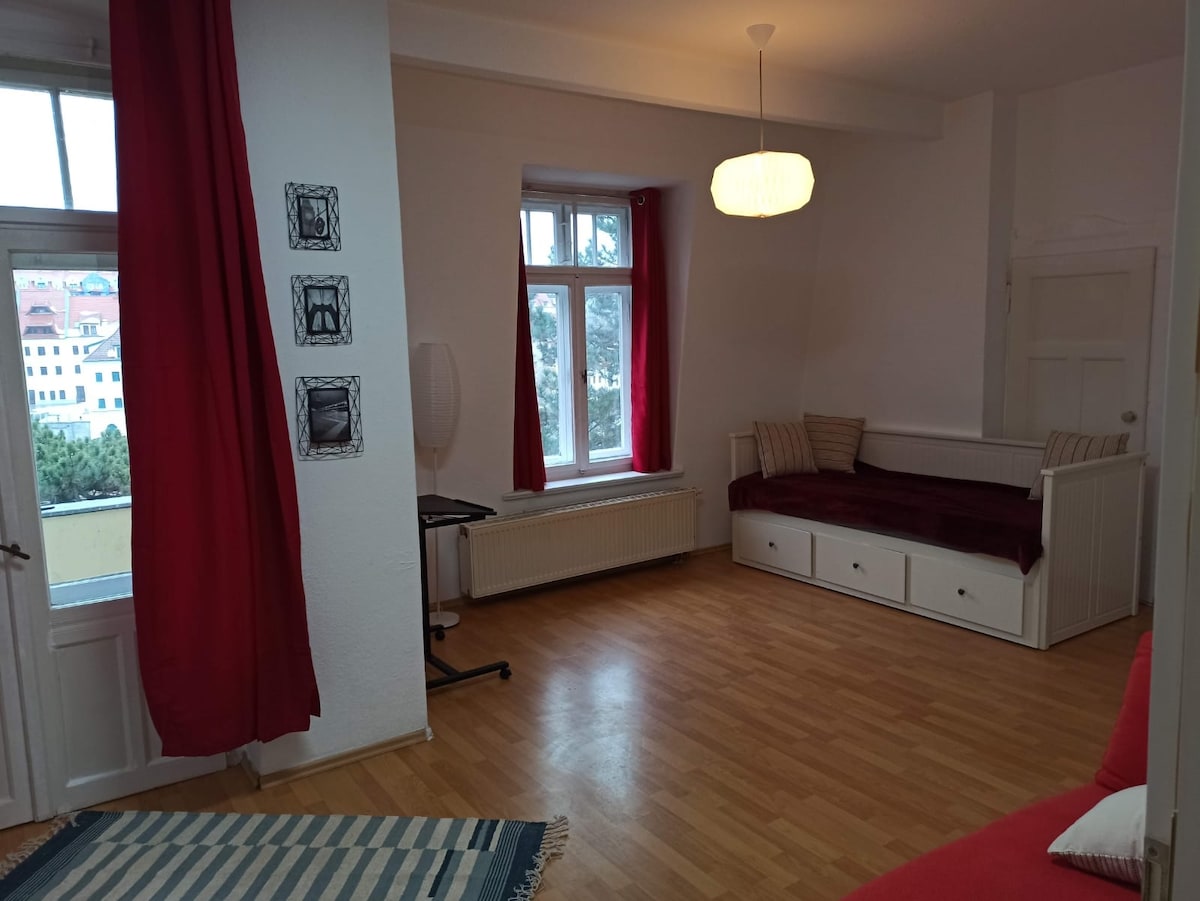 A spacious living area is enhanced by large windows allowing natural light to fill the space. Two red curtains frame the windows, while a light-colored rug lies on the wooden floor. A seating area with cushions is visible, alongside a minimalist lamp and a small table.