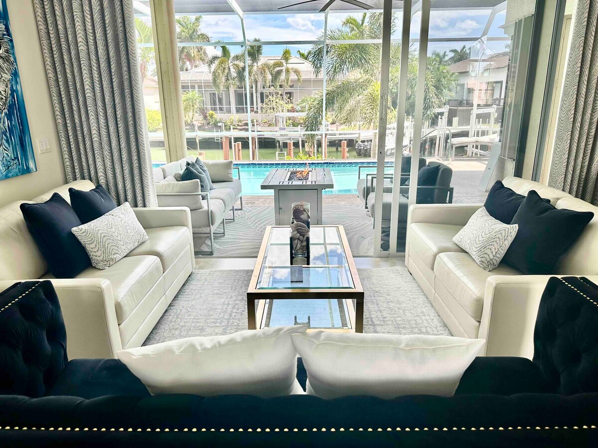 The living area features a pair of stylish white sofas accented with dark throw pillows, arranged around a glass coffee table. Large windows provide a view of the heated pool and lush palm trees, enhancing the space with natural light.