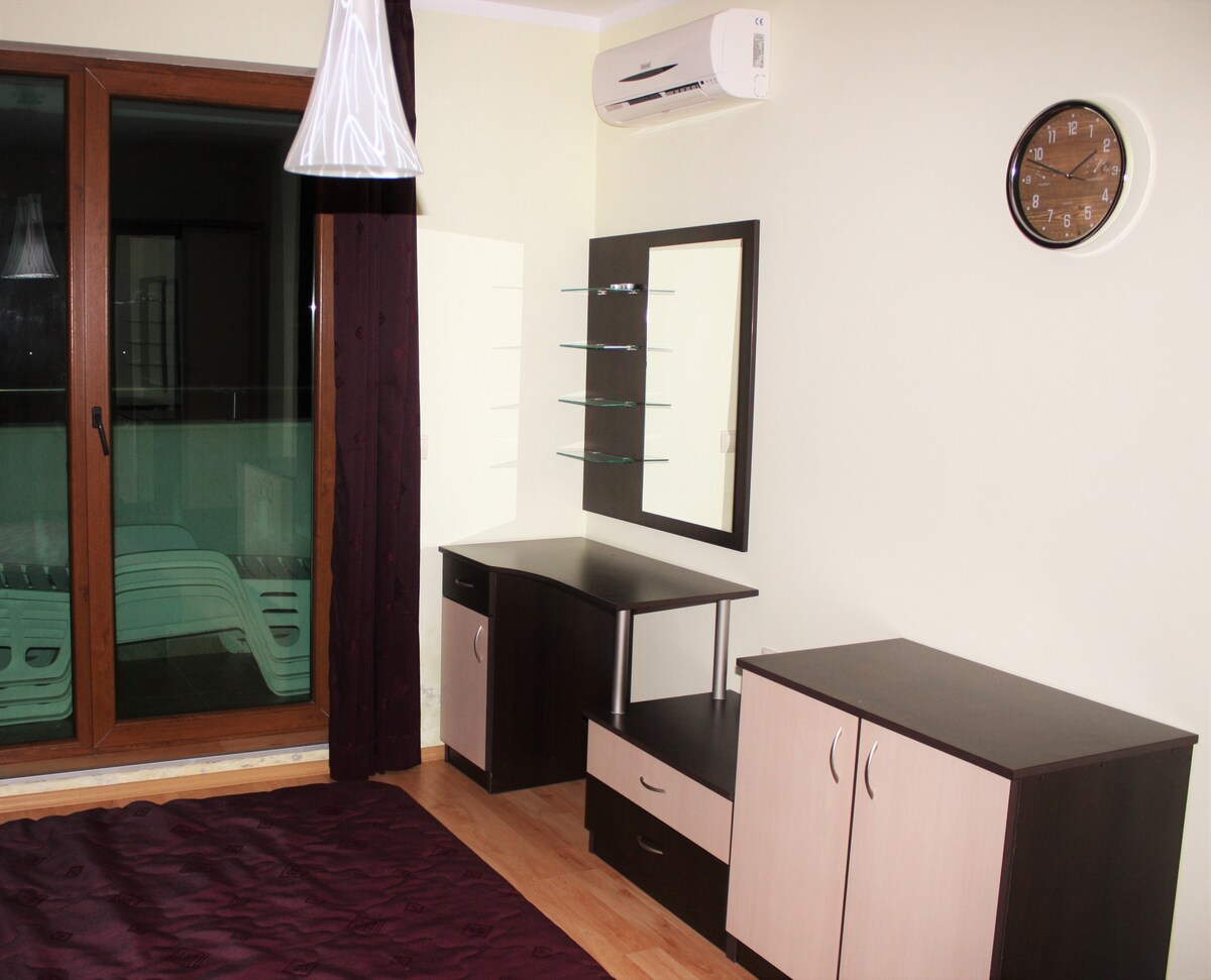 A spacious bedroom features a dark wooden floor and light walls. A stylish desk with a mirror and shelving unit is positioned beside a low wooden cabinet. A wall clock is mounted above, while a large window reveals a balcony with lounge chairs outside.