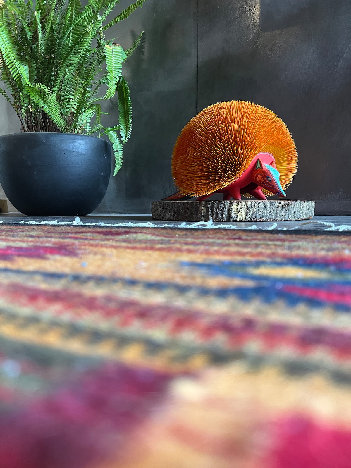 A vibrant Alebrije sculpture featuring bright orange and red colors is displayed on a wooden base. A textured rug in varied hues provides a warm foundation beneath it. In the background, a potted fern adds a touch of greenery, complementing the artistic decor.