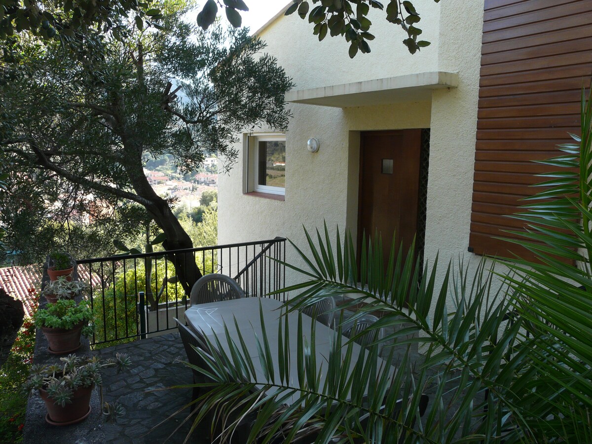 A private terrace is surrounded by lush greenery and potted plants. A table with chairs is situated on the stone patio, offering a peaceful setting. The entrance to the building features a wooden door, with a nearby window allowing natural light to enter.