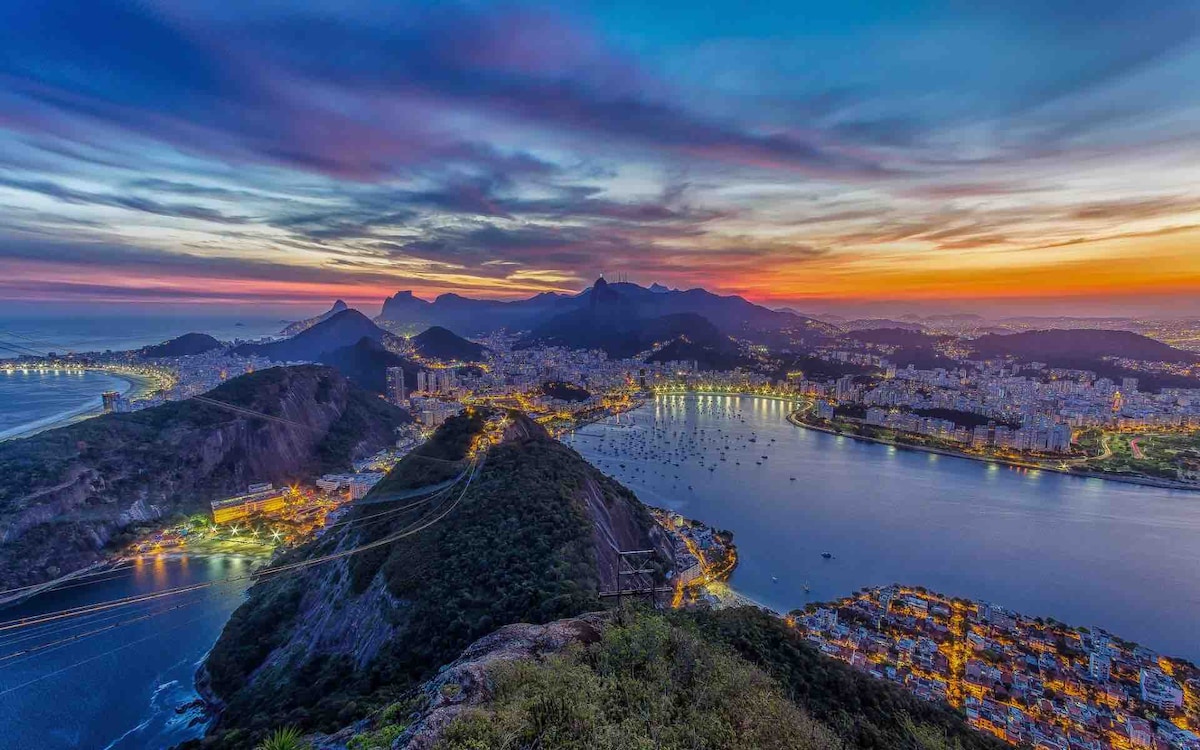 A sweeping view of Rio de Janeiro at sunset, showcasing the coastline and hills. The scene is illuminated by city lights reflecting on the water, while a vibrant sky transitions from orange to deep blue, framing the iconic Sugar Loaf Mountain and surrounding landscape.