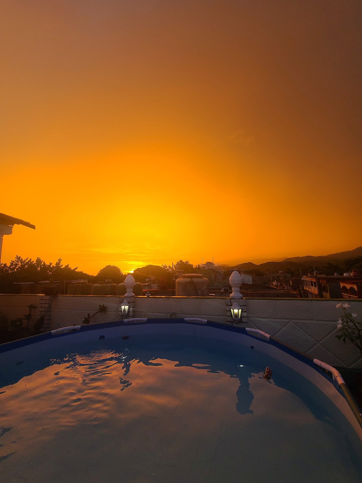 A rooftop terrace features a swimming pool reflecting the orange hues of a sunset. Soft lighting from decorative lamps illuminates the surrounding area. The landscape includes distant mountain silhouettes and rooftops, creating a serene backdrop for relaxation.
