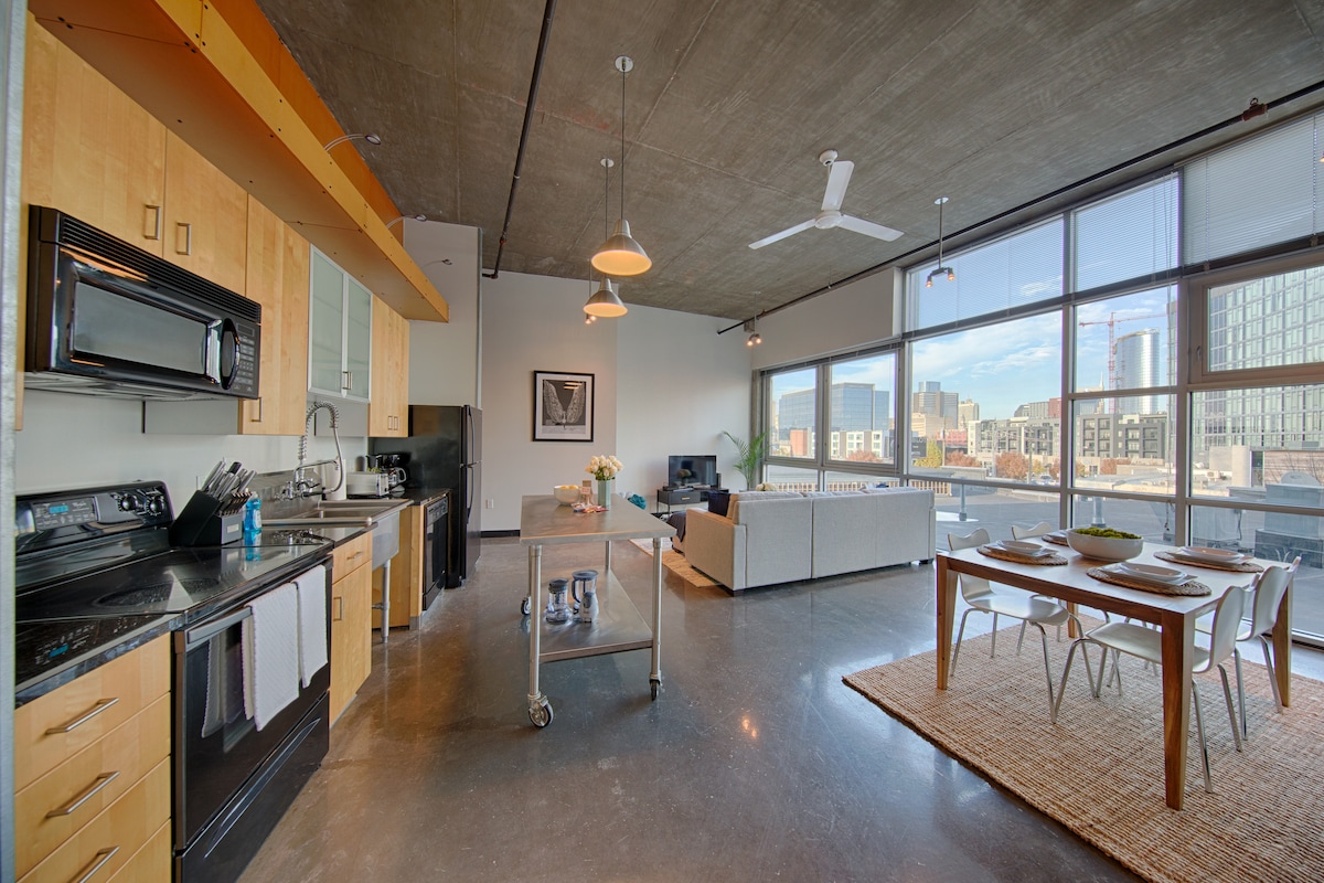 An open-concept kitchen features wooden cabinets and modern appliances, including a stove and microwave. A dining area with a wooden table is complemented by stylish place settings. Large windows offer views of the city skyline, allowing natural light to fill the space.