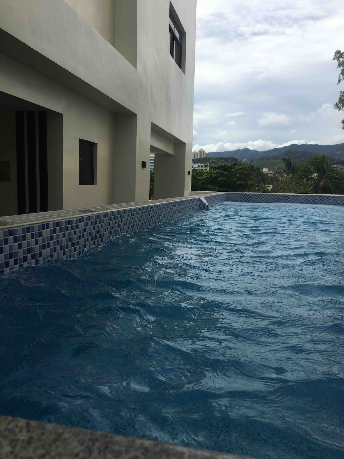 A pool area is showcased, featuring clear blue water reflecting the sky. The tile border contrasts with the smooth pool surface. Lush greenery and hills are visible in the background, enhancing the serene setting. The structure above presents several windows, allowing natural light to enter.
