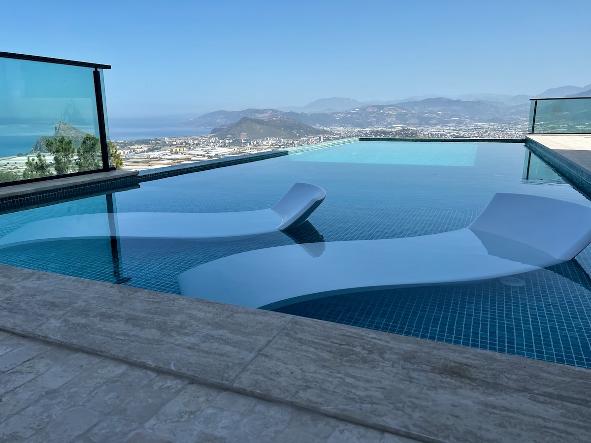 An infinity pool is distinctly visible, reflecting the surrounding landscape, with lounge chairs partially submerged at each end. Clear blue water stretches towards the horizon, showcasing panoramic views of the Mediterranean and distant hills, accented by a smooth stone border.
