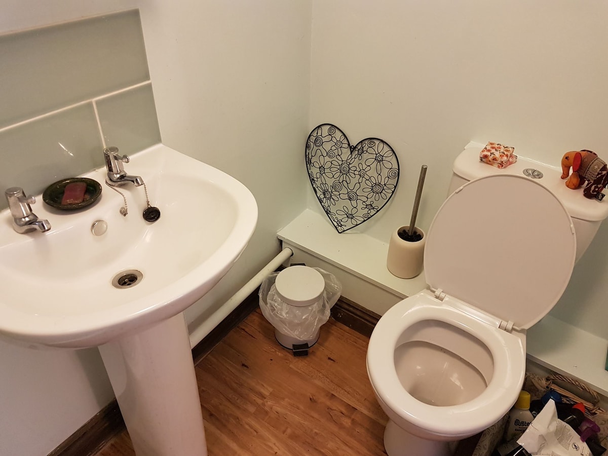 A compact en suite bathroom features a white pedestal sink and a toilet. On the wall, a decorative heart-shaped piece is displayed, contributing a personal touch. The floor is finished in wood, and a wastebasket is situated beside the sink.