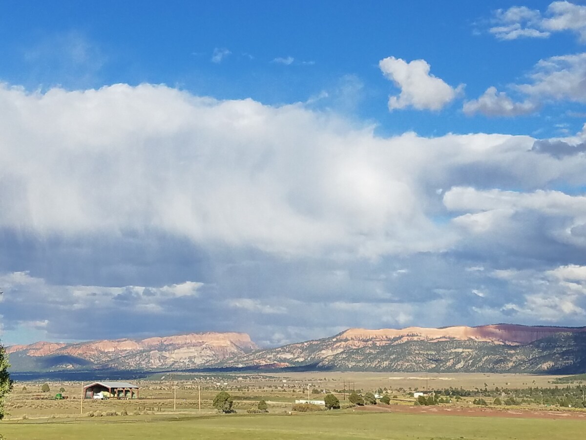 Wide open spaces are framed by distant mountains under a blue sky adorned with fluffy white clouds. The stunning layered cliffs display light and shadow, reflecting the natural beauty of Garfield County's landscape.
