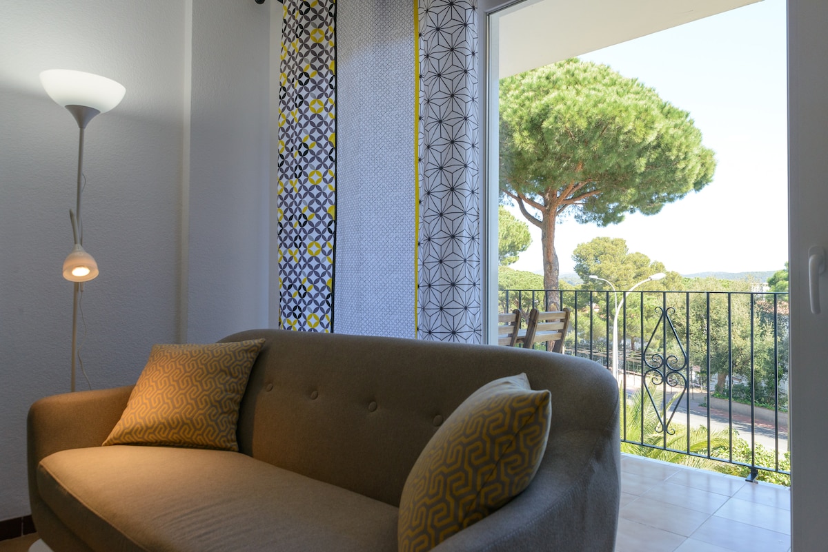 A comfortable sofa is positioned near a window with patterned curtains, allowing natural light to fill the space. A view of lush greenery and a balcony with outdoor seating is visible beyond the glass door, adding an element of serenity to the living area.