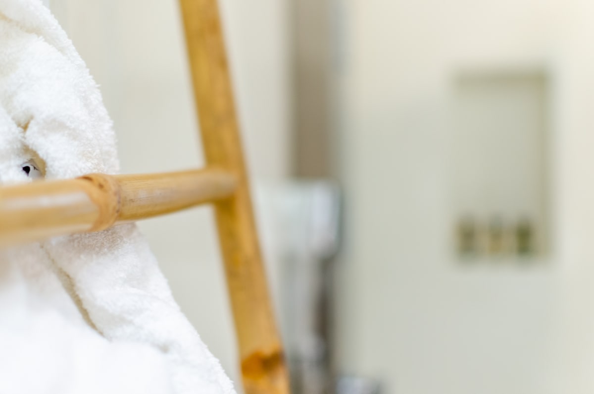 A bamboo ladder holds a stack of fluffy white towels, creating an organized and serene bathroom space. The background features soft neutral tones, with bathroom essentials subtly visible, accentuating a clean and functional design.