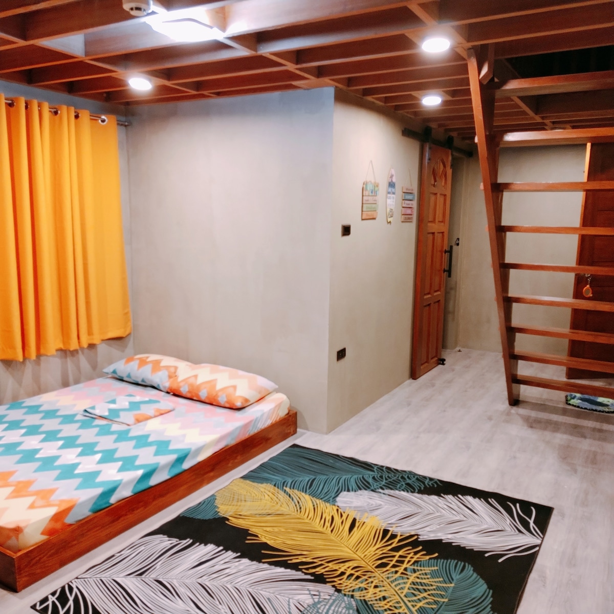 A bright room features a spacious bed with a colorful patterned comforter. Bright orange curtains frame the window, while a lively area rug with leaf designs adds a touch of color to the space. A wooden ladder leads to an elevated area.