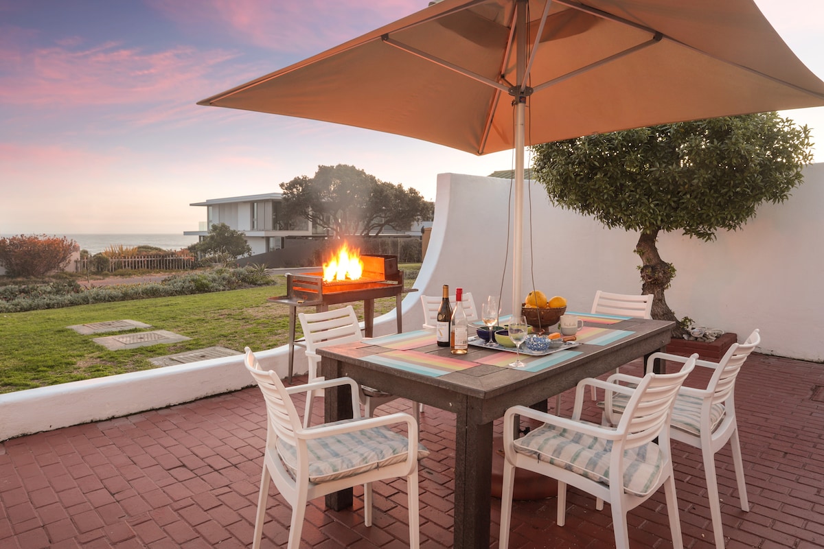 An outdoor patio area is highlighted by a dining table surrounded by six white chairs. A shaded umbrella provides cover over the table, which is set with plates, glasses, and fruit. A braai grill features a glowing flame, with a landscaped area visible in the background.