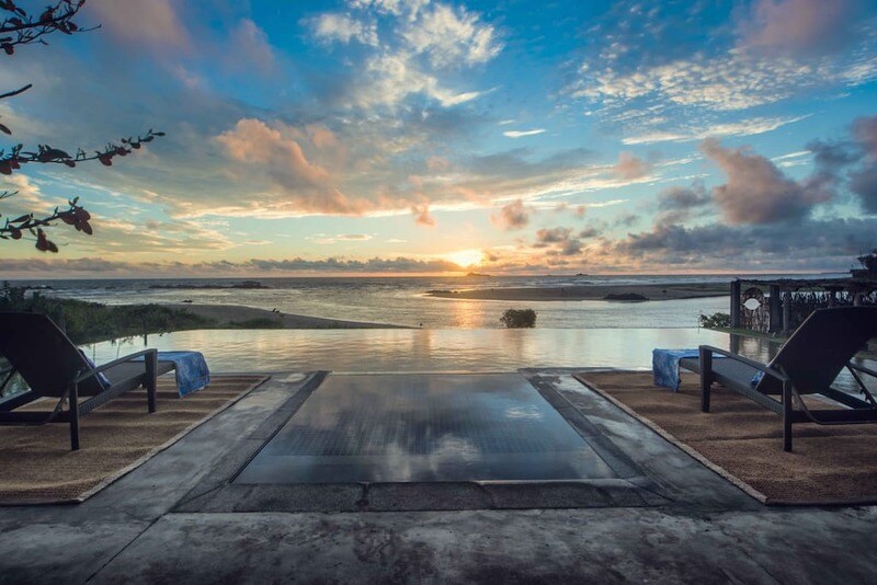 Two lounge chairs sit beside an infinity pool, overlooking a tranquil ocean at sunset. The sky is painted with soft hues of orange and blue, while the horizon reveals silhouettes of distant landmasses reflecting in the water.
