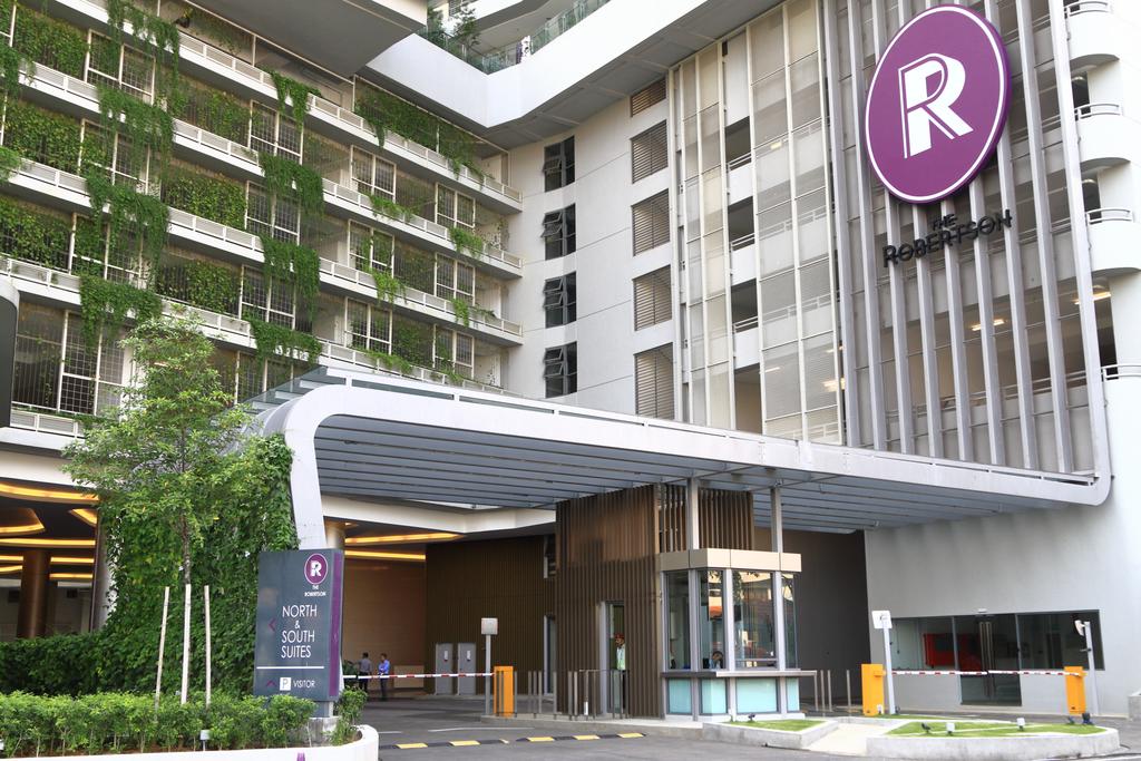 The entrance of the serviced apartment complex is marked by a modern façade featuring lush greenery and sleek architectural lines. The prominently displayed logo adds branding to the building. A covered driveway provides access, enhancing convenience for guests arriving by vehicle.