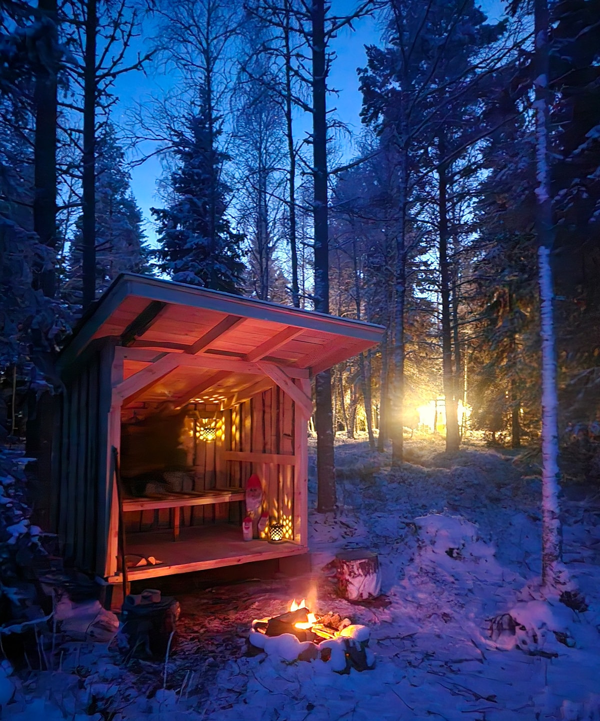 An outdoor shelter is illuminated by warm lights, nestled among tall pine trees in a snowy forest. A small campfire burns in front, surrounded by logs. The twilight sky adds a peaceful ambiance, while distant lights hint at nearby surroundings.
