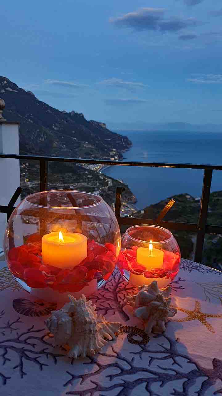 A serene balcony setting is presented during twilight, featuring two glass candle holders surrounded by rose petals. A gentle glow emanates from the candles, reflecting off the ocean view and illuminated hills in the background. Decorative seashells rest on a patterned tablecloth.