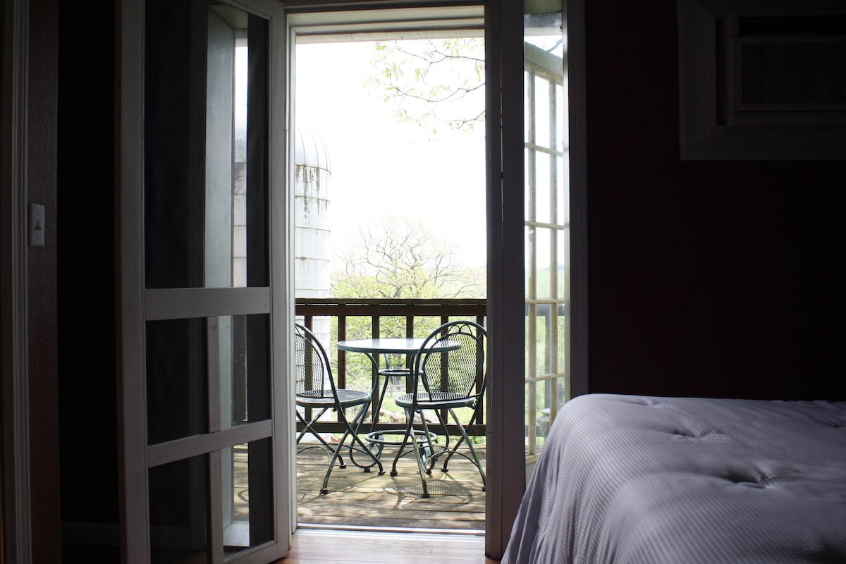 A view from the suite showcases an outdoor balcony with two metal chairs and a small table overlooking greenery. An open sliding door allows light to enter the room, which features a spacious bed and neutral-toned bedding.