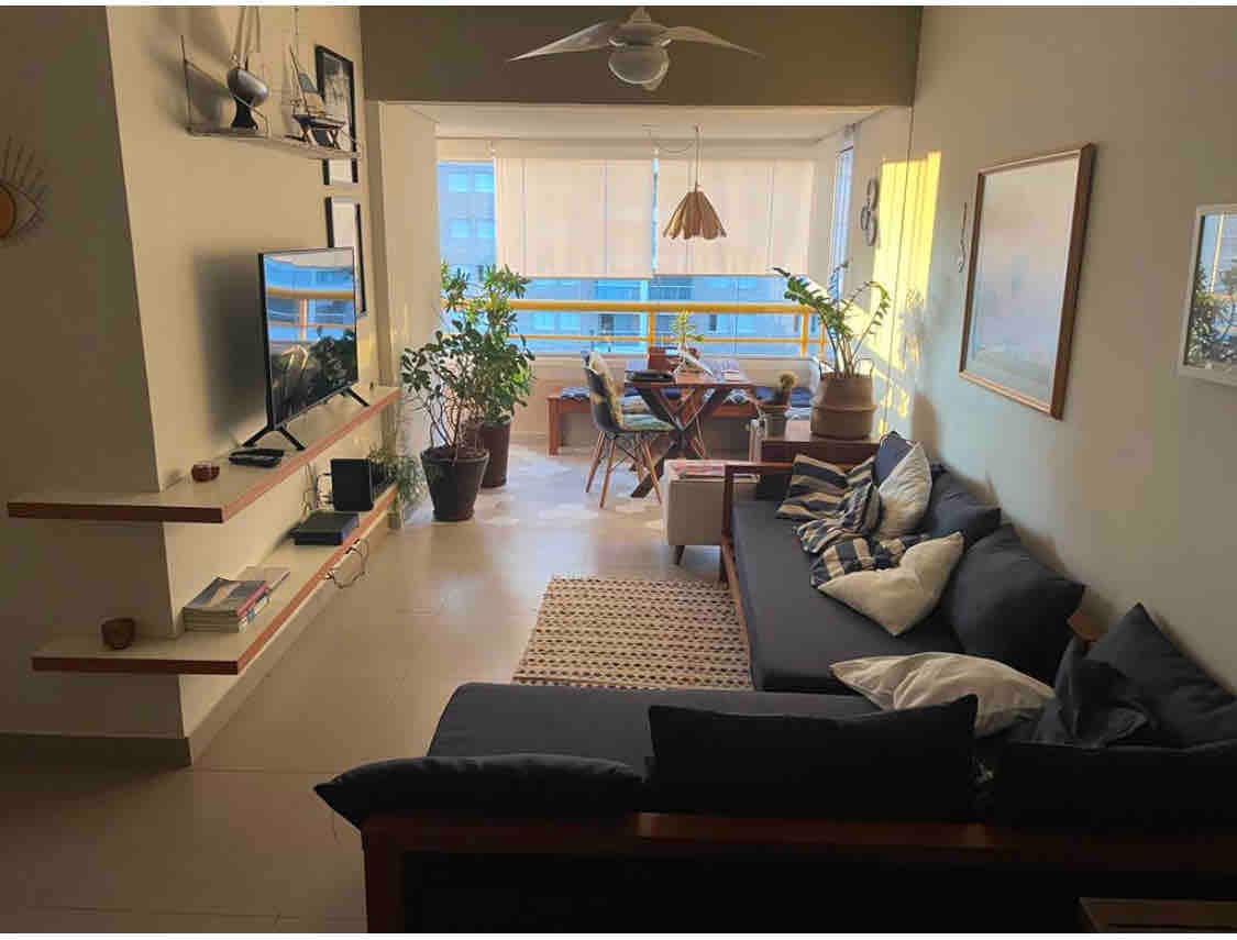 An inviting living space features a dark sectional sofa adorned with patterned cushions. A television is mounted on the wall, alongside a floating shelf. A dining area with wooden chairs is visible in the background, illuminated by natural light streaming through large windows.