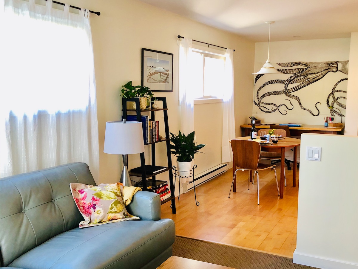 A cozy living area features a leather sofa adorned with a decorative pillow. A bookshelf stands nearby with greenery, while a dining table is visible in the background, complemented by a wall art piece of an octopus. Natural light enters through the windows.