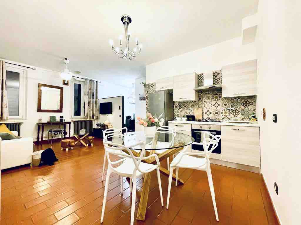 A modern kitchen and dining area are displayed, featuring a round wooden table surrounded by four white chairs. A stylish chandelier hangs above, and patterned tiles accentuate the kitchen's backsplash. A relaxation area with a light sofa is visible in the background.
