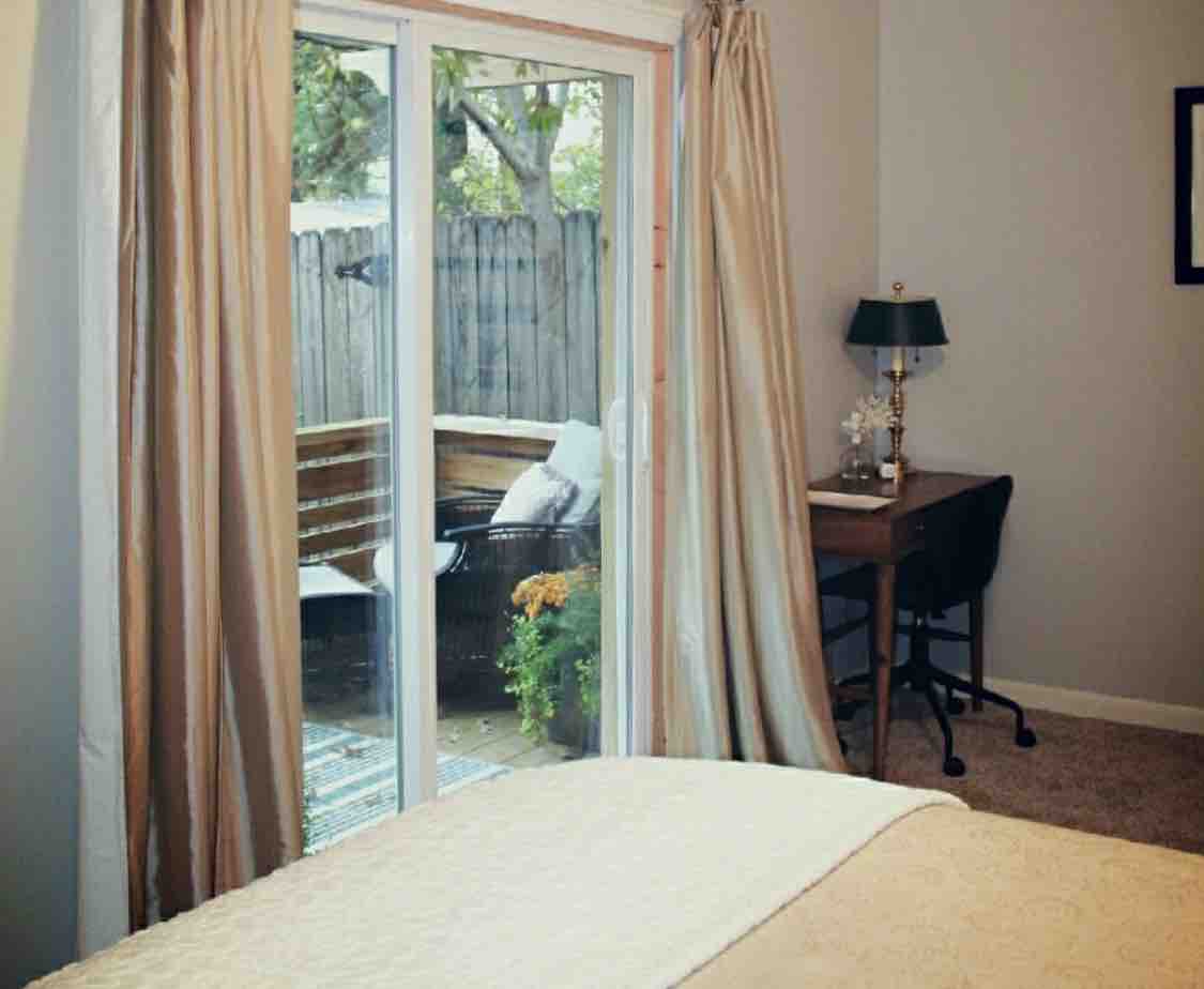 Natural light floods the room through sliding glass doors leading to a private deck. A cozy workspace with a chair and small table is positioned nearby, complemented by soft curtains that frame the entrance. A serene atmosphere is created by the neutral tones and outdoor greenery.