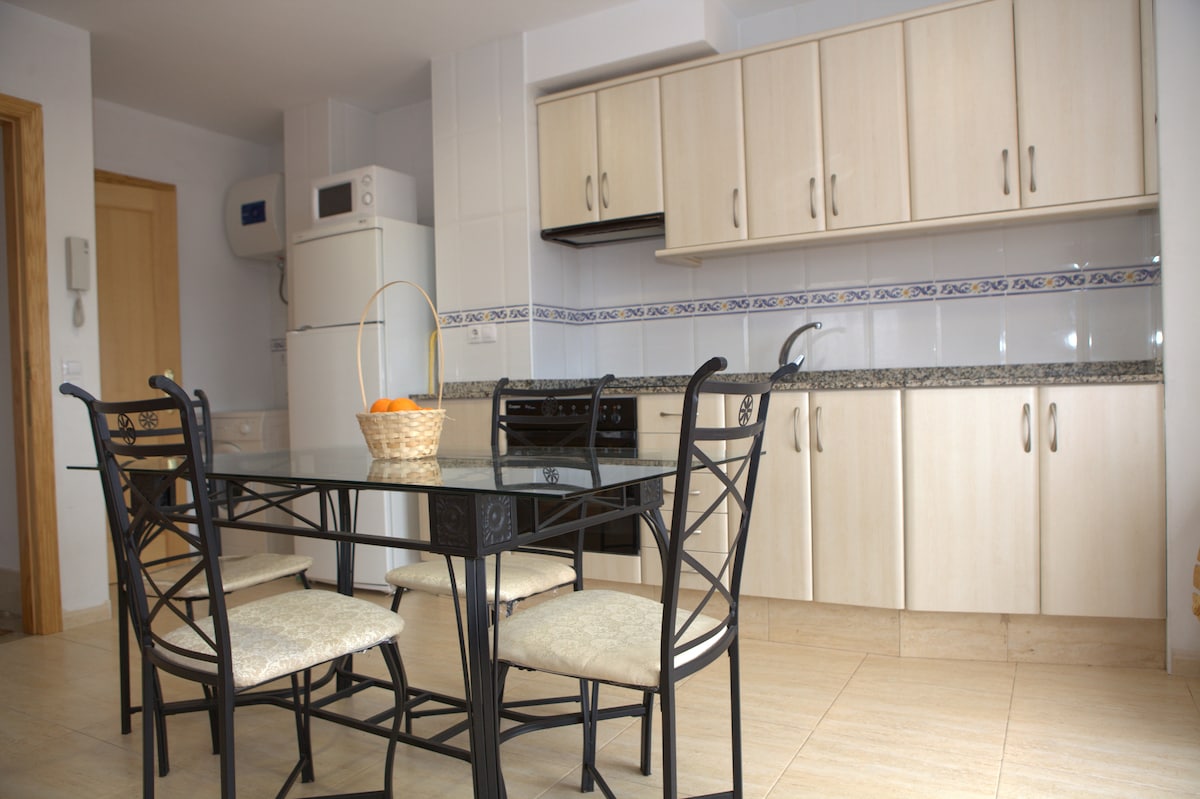 A dining area is shown, featuring a glass-top table surrounded by four black metal chairs. A sleek kitchen with light wood cabinets and granite countertops is visible in the background, complemented by white appliances. A basket of oranges is placed on the table.