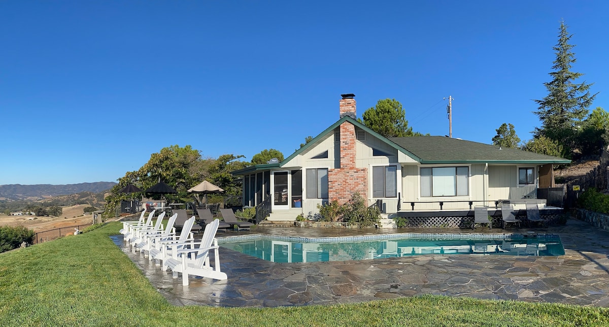 A spacious house with a green roof and brick chimney sits beside a private pool. An expansive patio area features white lounge chairs and umbrellas, providing comfortable spaces for relaxation. Lush grass and scenic views of the surrounding hills enhance the tranquil outdoor setting.