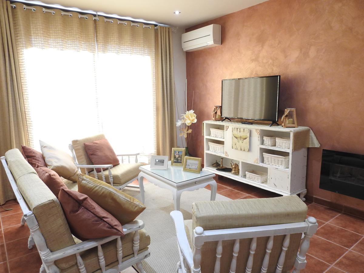 A bright living area features a cozy seating arrangement with neutral-toned sofas and decorative pillows. A white coffee table sits in the center, complemented by a media console displaying framed photos and decorative items. Natural light filters in through sheer curtains.