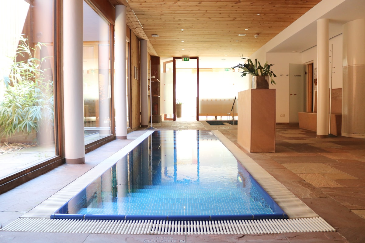 An indoor wellness area features a sleek swimming pool surrounded by natural stone flooring. Large glass doors allow natural light to fill the space, showcasing a warm wood ceiling. Decorative plants are positioned near the entrance, enhancing the tranquil atmosphere.