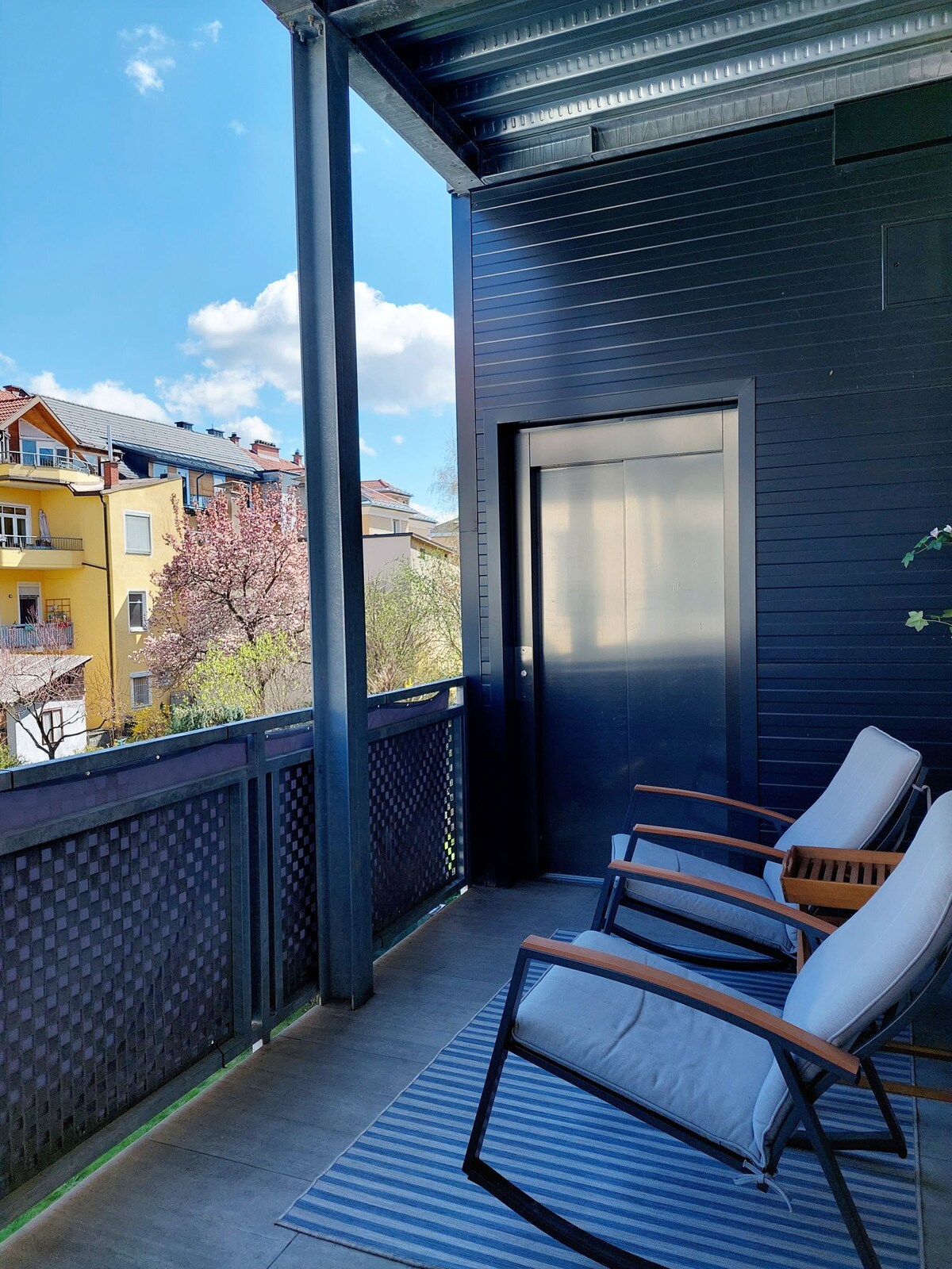 A spacious loggia offers seating with two comfortable chairs, positioned on a striped outdoor rug. The view includes neighboring buildings and greenery, illuminated by natural light under a partly cloudy sky.