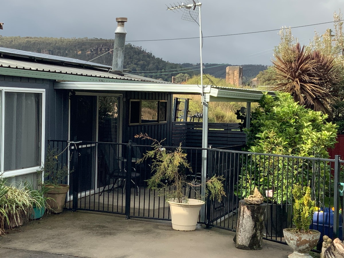 A fenced patio area is shown with a mixture of plants in pots and garden features. The entrance to the cabin is visible, along with a sturdy railing. The backdrop includes distant mountains, enhancing the natural setting.