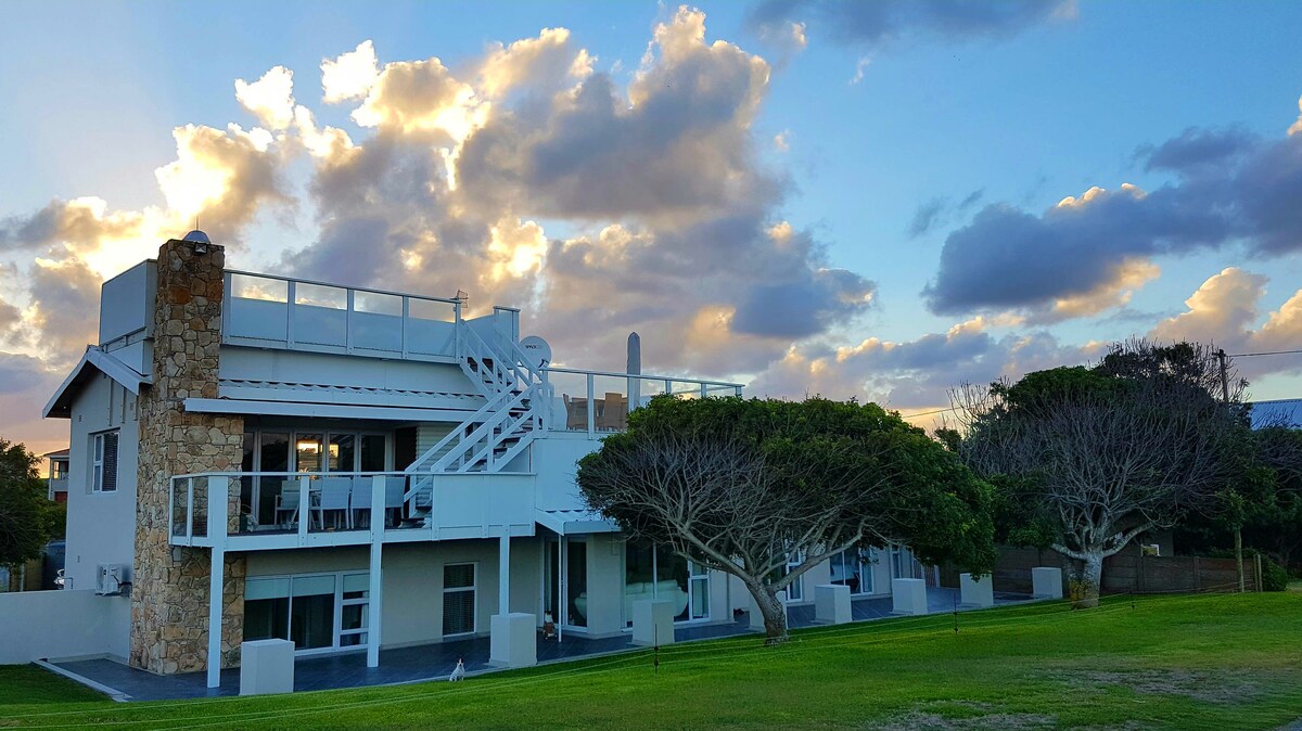 A modern two-story building is displayed against a backdrop of partly cloudy skies. The structure features a spacious deck, large windows, and a staircase leading to the upper level. Lush green grass surrounds the property, contributing to the serene atmosphere.