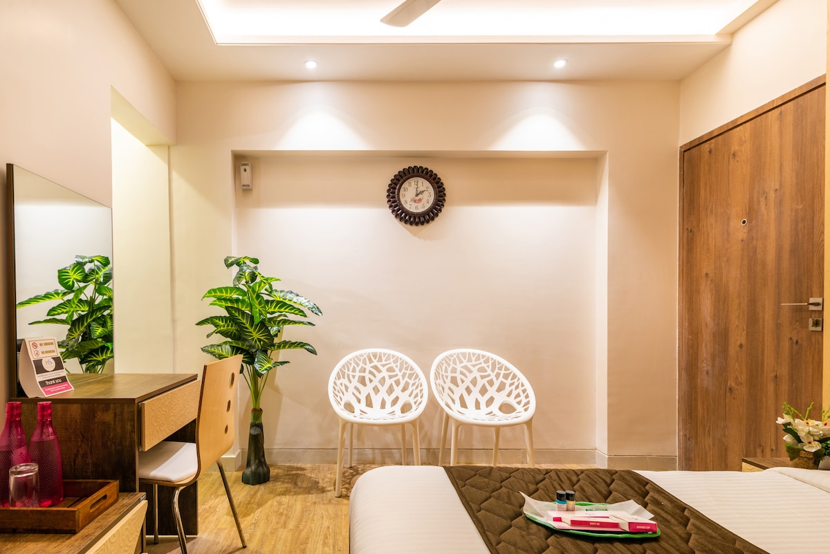 The studio features a modern design with a spacious bed at the forefront. Two white chairs are positioned against the wall, accompanied by a decorative plant. A wall clock and minimalistic mirror enhance the overall aesthetic, while soft lighting creates a comfortable ambiance.
